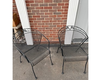 May include: Two black metal outdoor chairs with mesh seats and backs. The chairs have curved backs with decorative leaf accents. The chairs are on a concrete surface in front of a brick wall.