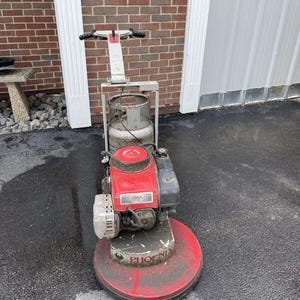 May include: A red and white gas-powered floor polisher with a white handle and a black engine. The engine has the text "Titan" and "OHV" on it. The polisher is sitting on a black asphalt surface. The polisher has a red circular base with the word "Phoenix" on it.