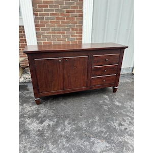 May include: A dark brown wooden cabinet with two doors and three drawers. The cabinet has a rectangular shape with a flat top and sits on short legs with silver accents. The wood grain is visible, and the hardware is a dark metal.
