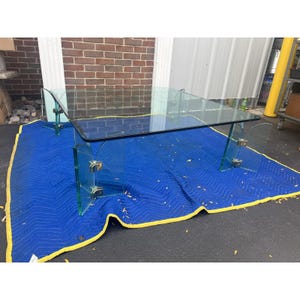 May include: A clear glass coffee table with a rectangular top and curved edges. The table is supported by clear glass legs with metal accents. The table is on a blue moving blanket with a yellow border.