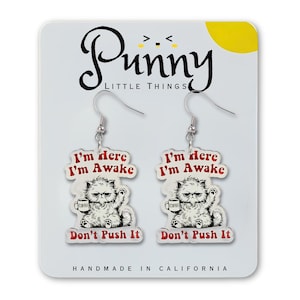 May include: A pair of novelty earrings featuring a grumpy cat design. Each earring displays a white cat holding a coffee cup, with the text "I'm here, I'm awake, Don't Push It" in red. The earrings are on a white card.