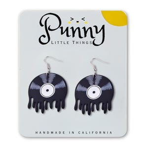 以下が含まれることがあります： 溶けたデザインの黒いビニールレコードのイヤリングのペア。イヤリングはシルバーのフックが付いており、「Punny Little Things」と「Handmade in California」の文字が入った白いカードに表示されています。