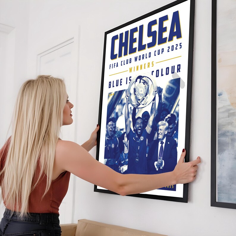 May include: A framed poster with the word "CHELSEA" in blue and white text. The poster commemorates the FIFA Club World Cup 2025 winners. The image features a team holding a trophy. The poster is being held by a person.