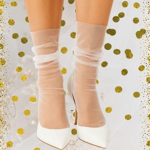 Op de afbeelding: Een paar witte pumps met spitse tenen. De hakken worden gecombineerd met doorschijnende, lichtgekleurde sokken. De achtergrond is wit met gouden confetti.