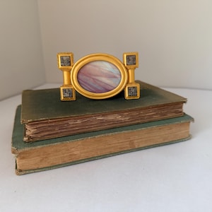 May include: A gold-toned metal belt buckle featuring a central oval inlay with swirling purple and white patterns. The buckle has a rectangular frame on each side, each containing four small square inlays with detailed black and white illustrations.