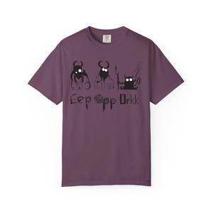 Può includere: T-shirt color prugna con grafica nera di tre creature dei cartoni animati e la scritta "Eep Opp Orkk". Le creature hanno corna e tengono in mano vari oggetti. La maglietta è realizzata in materiale morbido.