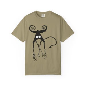 May include: A khaki-colored t-shirt featuring a black, stylized creature design. The creature has a head with two curled horns, two white eyes, and a long tail with an arrow-shaped tip. The design is centered on the front of the shirt.