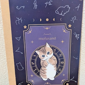 May include: A notebook with a navy blue cover featuring a cartoon cat illustration. The cover includes zodiac symbols, constellations, and the text "mofusand". The notebook has a tan spine.