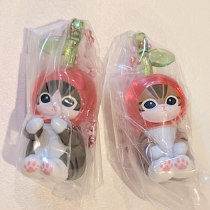 May include: Two cat figurines wearing red cherry-shaped hoods with green stems, each enclosed in clear plastic packaging. The cats have white and brown or black fur patterns, large dark eyes, and white paws with small pink paw pad details on the bottom.