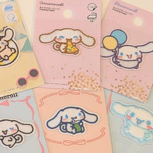 Peut inclure: Six écussons représentant le personnage Cinnamoroll sur des cartes de support individuelles. Les écussons brodés montrent le chiot blanc dans diverses positions, notamment avec des lunettes de soleil, une part de pizza, des ballons et une tasse.
