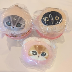 以下が含まれることがあります： 透明なプラスチック包装に入った3つの猫のぬいぐるみ。左上のぬいぐるみはグレーのトラ猫の顔、右上のぬいぐるみは金色の目をした黒い顔、下のぬいぐるみはピンクのフードを被った茶色と白の顔をしています。