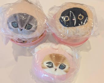 May include: Three plush cat toys, each encased in clear plastic. The cats have detailed faces and are wearing hooded outfits in shades of pink, tan, and black. The toys are in clear, pink-rimmed containers.