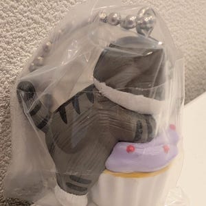 K&ouml;nnte beinhalten: Graue Katzenfigur mit schwarzen Streifen, die auf einem Cupcake mit lila Zuckerguss und roten Streuseln sitzt. Die Katze tr&auml;gt einen wei&szlig;en Kragen. Sie ist an einem silbernen Schl&uuml;sselanh&auml;nger befestigt.