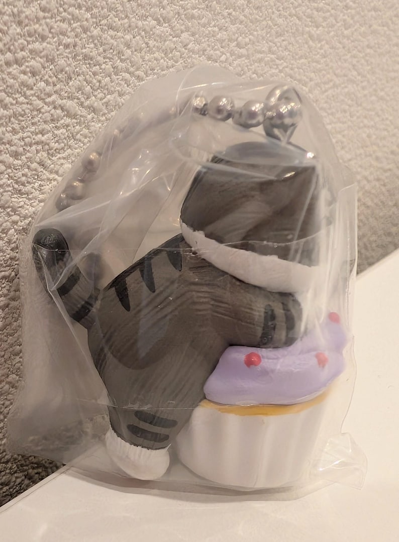 K&ouml;nnte beinhalten: Graue Katzenfigur mit schwarzen Streifen, die auf einem Cupcake mit lila Zuckerguss und roten Streuseln sitzt. Die Katze tr&auml;gt einen wei&szlig;en Kragen. Sie ist an einem silbernen Schl&uuml;sselanh&auml;nger befestigt.