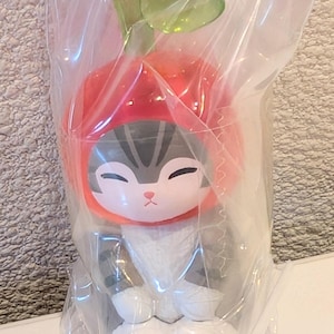 May include: A small cat figurine wearing a red apple-shaped hat with a green stem and leaf on top. The cat has gray and white fur, closed eyes, and white paws with pink paw pads. The entire figure is encased in a clear, crinkled plastic bag.