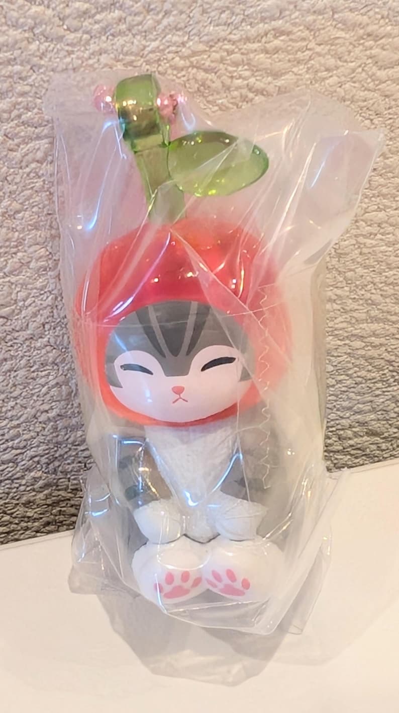 May include: A small cat figurine wearing a red apple-shaped hat with a green stem and leaf on top. The cat has gray and white fur, closed eyes, and white paws with pink paw pads. The entire figure is encased in a clear, crinkled plastic bag.