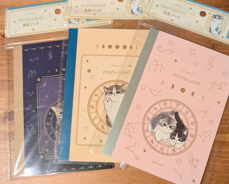 May include: A collection of A6 notebooks featuring cat illustrations and zodiac-themed designs. The notebooks are in various colors, including navy, peach, and pink, with the brand name "mofusand" printed on the covers.