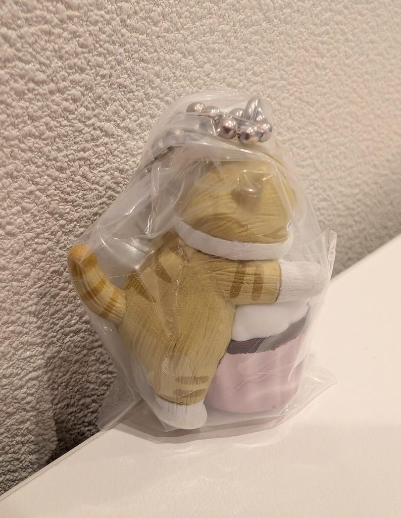K&ouml;nnte beinhalten: Ein katzenf&ouml;rmiger Schl&uuml;sselanh&auml;nger, verpackt in durchsichtigem Kunststoff. Die Katze ist hellbraun mit braunen Streifen und wei&szlig;en Akzenten und umarmt einen rosa-wei&szlig;en Cupcake. Der Schl&uuml;sselanh&auml;nger hat einen silberfarbenen Ring.