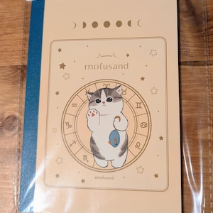 May include: A notebook with a beige cover featuring a cartoon cat holding a fish, surrounded by zodiac symbols and stars. The text "mofusand" is above the cat. The spine is blue.