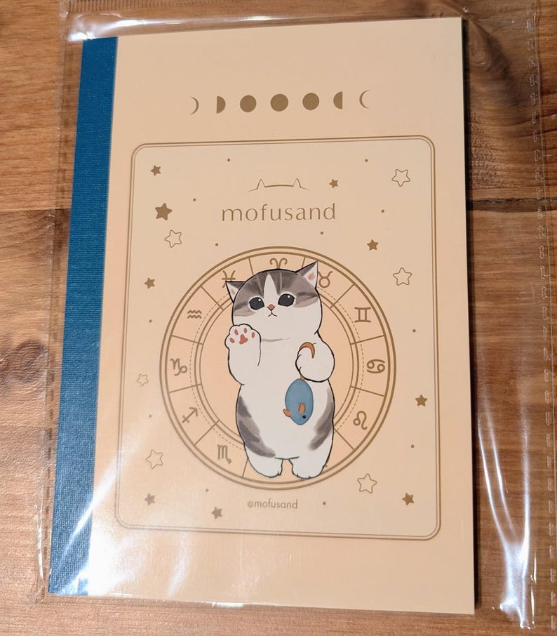 May include: A notebook with a beige cover featuring a cartoon cat holding a fish, surrounded by zodiac symbols and stars. The text "mofusand" is above the cat. The spine is blue.