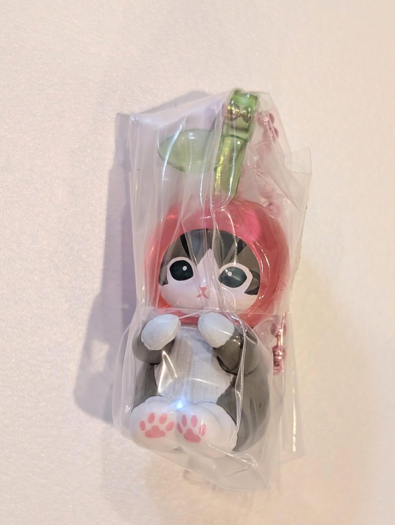 May include: A small cat figurine wearing a pink apple-shaped hood with a green leaf on top, encased in a clear plastic bag. The cat has black and white fur, large dark eyes, and visible pink paw pads on its front feet, attached to a pink chain.