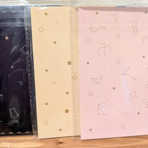 May include: Three stationery notebooks in a clear plastic sleeve. The notebooks have different colored backgrounds: navy blue, light beige, and pink. Each notebook features a constellation and star pattern in gold.