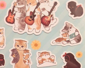 May include: A collection of die-cut stickers featuring illustrated cats in various poses, including three cats playing guitars, a cat holding two smaller kittens, a cat looking at a phone, a sleeping cat, and black cats with red bows.