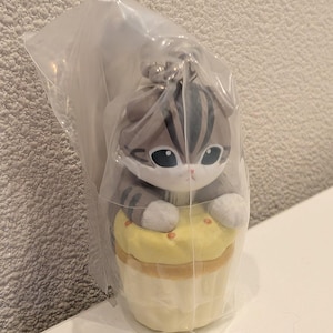 K&ouml;nnte beinhalten: Pl&uuml;schkatzenfigur mit grau-wei&szlig;en Streifen, gro&szlig;en Augen und wei&szlig;en Pfoten, die auf einem gelben Cupcake sitzt. Die Figur befindet sich in einer durchsichtigen Plastikt&uuml;te. Der Cupcake hat einen hellbraunen Boden und kleine orangefarbene Streusel.