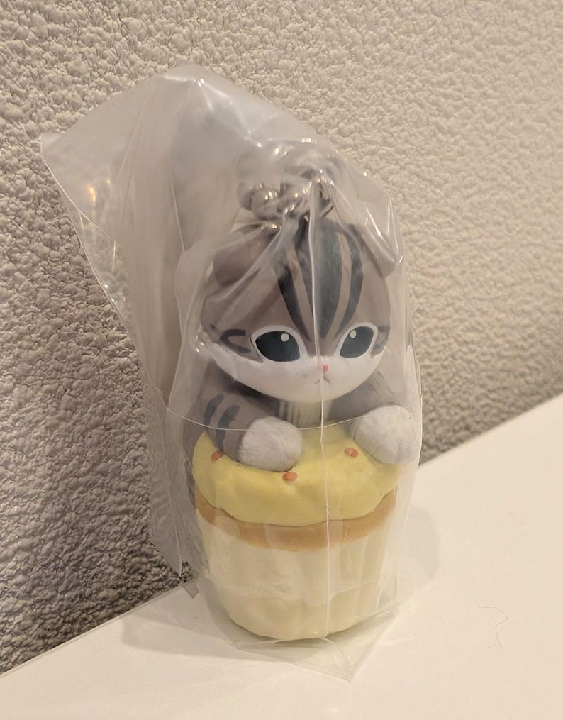 K&ouml;nnte beinhalten: Pl&uuml;schkatzenfigur mit grau-wei&szlig;en Streifen, gro&szlig;en Augen und wei&szlig;en Pfoten, die auf einem gelben Cupcake sitzt. Die Figur befindet sich in einer durchsichtigen Plastikt&uuml;te. Der Cupcake hat einen hellbraunen Boden und kleine orangefarbene Streusel.