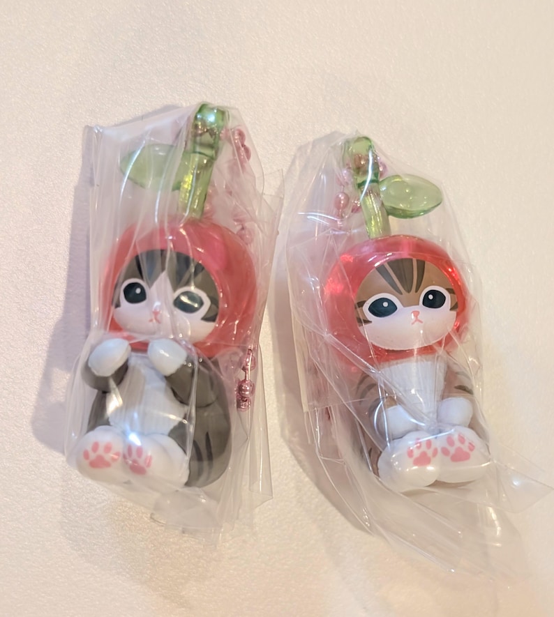 May include: Two cat figurines wearing red cherry-shaped hoods with green stems, each enclosed in clear plastic packaging. The cats have white and brown or black fur patterns, large dark eyes, and white paws with small pink paw pad details on the bottom.