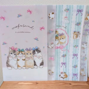 Peut inclure: Ensemble de chemises de classement en plastique transparent sur le thème des chats. Les chemises présentent un schéma de couleurs rose clair et bleu avec des illustrations de chatons, de nœuds et de motifs floraux. Le texte "mofusand" et "A cute little Nikukyu" est également présent.