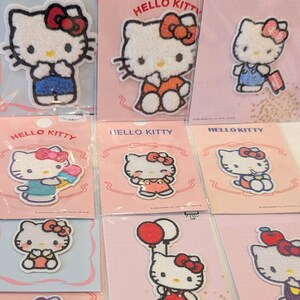 May include: A collection of embroidered Hello Kitty iron-on patches displayed on individual pink and white backing cards. Each patch features the white cat character in various outfits and poses, including holding balloons, ice cream, or luggage.