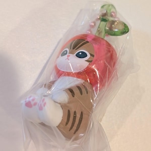 May include: A small brown tabby cat figurine wearing a red apple-shaped hood with a green leaf on top. The cat has white paws with pink paw pads and is encased in a clear plastic bag with a pink ball chain attached to the top of the apple stem.