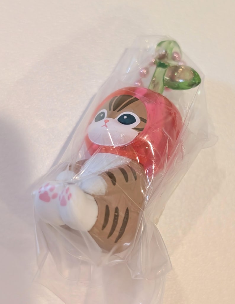 May include: A small brown tabby cat figurine wearing a red apple-shaped hood with a green leaf on top. The cat has white paws with pink paw pads and is encased in a clear plastic bag with a pink ball chain attached to the top of the apple stem.