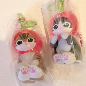 May include: Two cat figurines encased in clear plastic packaging, each wearing a red apple-shaped hood with a green leaf stem. The cats have large eyes, white paws with pink paw pads, and are attached to pink ball chain keychains for hanging.