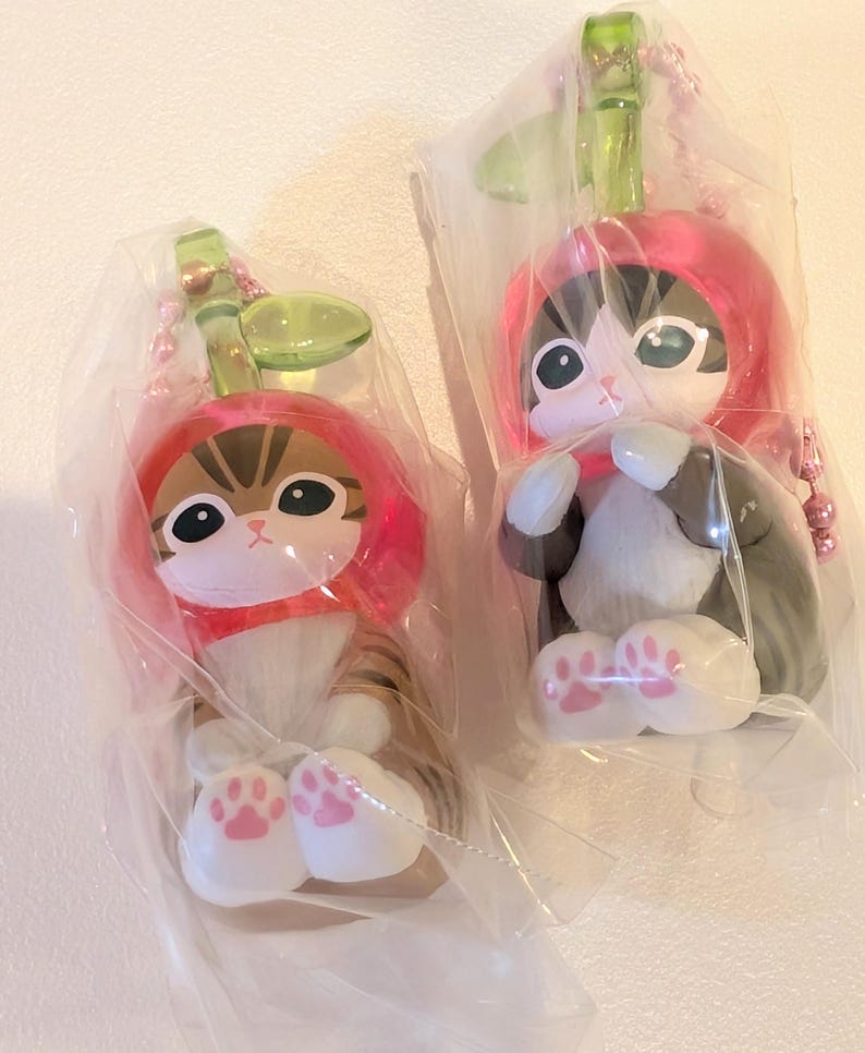 May include: Two cat figurines encased in clear plastic packaging, each wearing a red apple-shaped hood with a green leaf stem. The cats have large eyes, white paws with pink paw pads, and are attached to pink ball chain keychains for hanging.