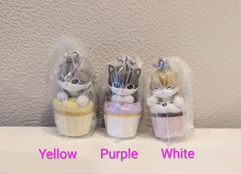 K&ouml;nnte beinhalten: Drei katzenf&ouml;rmige Figuren in Plastikverpackung. Jede Katze sitzt auf einem Cupcake. Die Cupcakes sind gelb, lila und wei&szlig;. Der Text "Yellow", "Purple" und "White" befindet sich darunter.