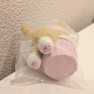 K&ouml;nnte beinhalten: Eine kleine, gelb-wei&szlig;e Katzenfigur mit rosa Pfotenballen ist teilweise in einer durchsichtigen Plastikt&uuml;te zu sehen. Die Katze ist mit dem Hinterteil zum Betrachter positioniert, neben einem rosa zylindrischen Objekt. Die Figur ist wahrscheinlich ein Sammlerst&uuml;ck oder ein Spielzeug.