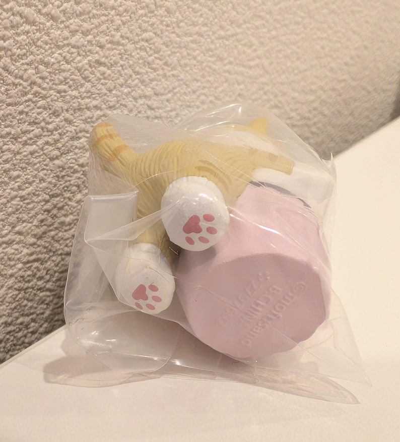 K&ouml;nnte beinhalten: Eine kleine, gelb-wei&szlig;e Katzenfigur mit rosa Pfotenballen ist teilweise in einer durchsichtigen Plastikt&uuml;te zu sehen. Die Katze ist mit dem Hinterteil zum Betrachter positioniert, neben einem rosa zylindrischen Objekt. Die Figur ist wahrscheinlich ein Sammlerst&uuml;ck oder ein Spielzeug.