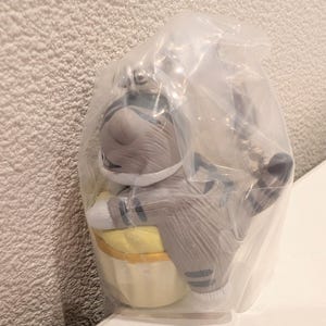 K&ouml;nnte beinhalten: Graue Katzenfigur in einer durchsichtigen Plastikt&uuml;te. Die Katze tr&auml;gt einen grau-wei&szlig;en Astronautenanzug und h&auml;lt einen gelben Gegenstand. Die Figur hat schwarze Streifen und ist an einem silbernen Schl&uuml;sselanh&auml;nger befestigt.