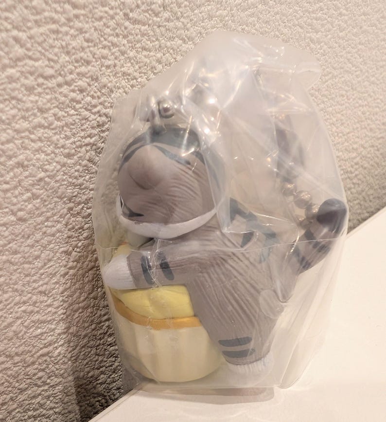 K&ouml;nnte beinhalten: Graue Katzenfigur in einer durchsichtigen Plastikt&uuml;te. Die Katze tr&auml;gt einen grau-wei&szlig;en Astronautenanzug und h&auml;lt einen gelben Gegenstand. Die Figur hat schwarze Streifen und ist an einem silbernen Schl&uuml;sselanh&auml;nger befestigt.
