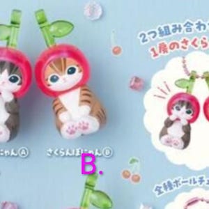 May include: Two small cat figurines, one dark brown and one light brown, each wearing a red cherry-shaped hood with a green stem and leaf. The text includes Japanese characters and the labels A and B. The figures feature large eyes and white paws.