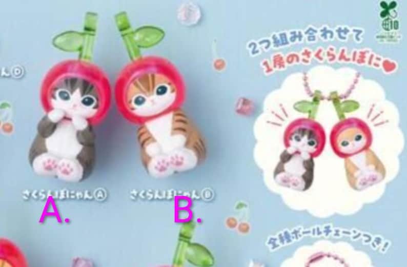 May include: Two small cat figurines, one dark brown and one light brown, each wearing a red cherry-shaped hood with a green stem and leaf. The text includes Japanese characters and the labels A and B. The figures feature large eyes and white paws.