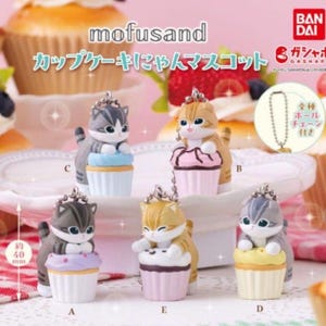 K&ouml;nnte beinhalten: Kleine Katzenfiguren auf Cupcake-Sockeln. Die Figuren, etwa 4 cm hoch, sind in verschiedenen Farben erh&auml;ltlich und haben eine Kette. Der Text "mofusand" und japanische Schriftzeichen sind sichtbar.