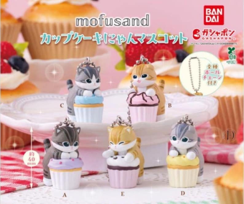 K&ouml;nnte beinhalten: Kleine Katzenfiguren auf Cupcake-Sockeln. Die Figuren, etwa 4 cm hoch, sind in verschiedenen Farben erh&auml;ltlich und haben eine Kette. Der Text "mofusand" und japanische Schriftzeichen sind sichtbar.