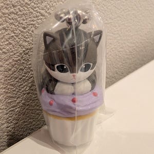 K&ouml;nnte beinhalten: Ein katzenf&ouml;rmiger Schl&uuml;sselanh&auml;nger-Figur in einer durchsichtigen Plastikt&uuml;te. Die Figur ist eine grau-wei&szlig;e Katze, die auf einem lila Cupcake mit rosa Streuseln sitzt. Der Cupcake steht auf einer wei&szlig;en Basis. Der Schl&uuml;sselanh&auml;nger hat einen silbernen Metallverschluss.