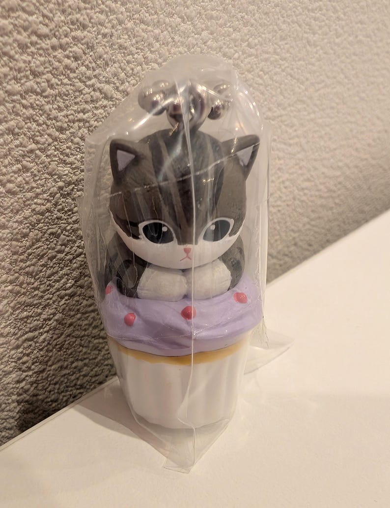 K&ouml;nnte beinhalten: Ein katzenf&ouml;rmiger Schl&uuml;sselanh&auml;nger-Figur in einer durchsichtigen Plastikt&uuml;te. Die Figur ist eine grau-wei&szlig;e Katze, die auf einem lila Cupcake mit rosa Streuseln sitzt. Der Cupcake steht auf einer wei&szlig;en Basis. Der Schl&uuml;sselanh&auml;nger hat einen silbernen Metallverschluss.