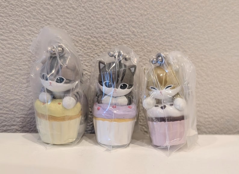 K&ouml;nnte beinhalten: Drei katzenf&ouml;rmige Figuren, jede auf einem Cupcake, in transparenter Kunststoffverpackung. Die Cupcakes sind gelb, lila und rosa. Die Katzen haben unterschiedliche Farben und Muster.