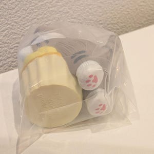 K&ouml;nnte beinhalten: Ein Pl&uuml;sch-Katzen-Spielzeug in einer durchsichtigen Plastikt&uuml;te. Das Spielzeug hat eine hellgelbe, cupcake-f&ouml;rmige Basis mit Text und einen grauen Katzenk&ouml;rper mit wei&szlig;en Pfoten und rosa Pfotenballen. Das Spielzeug ist f&uuml;r den Verkauf verpackt.