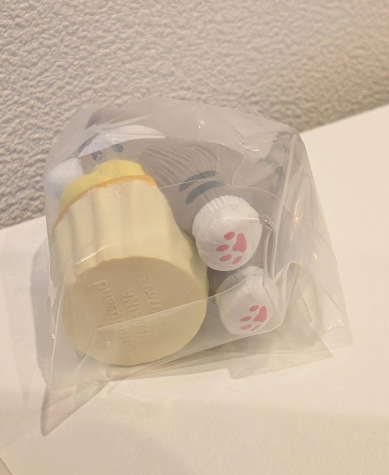 K&ouml;nnte beinhalten: Ein Pl&uuml;sch-Katzen-Spielzeug in einer durchsichtigen Plastikt&uuml;te. Das Spielzeug hat eine hellgelbe, cupcake-f&ouml;rmige Basis mit Text und einen grauen Katzenk&ouml;rper mit wei&szlig;en Pfoten und rosa Pfotenballen. Das Spielzeug ist f&uuml;r den Verkauf verpackt.